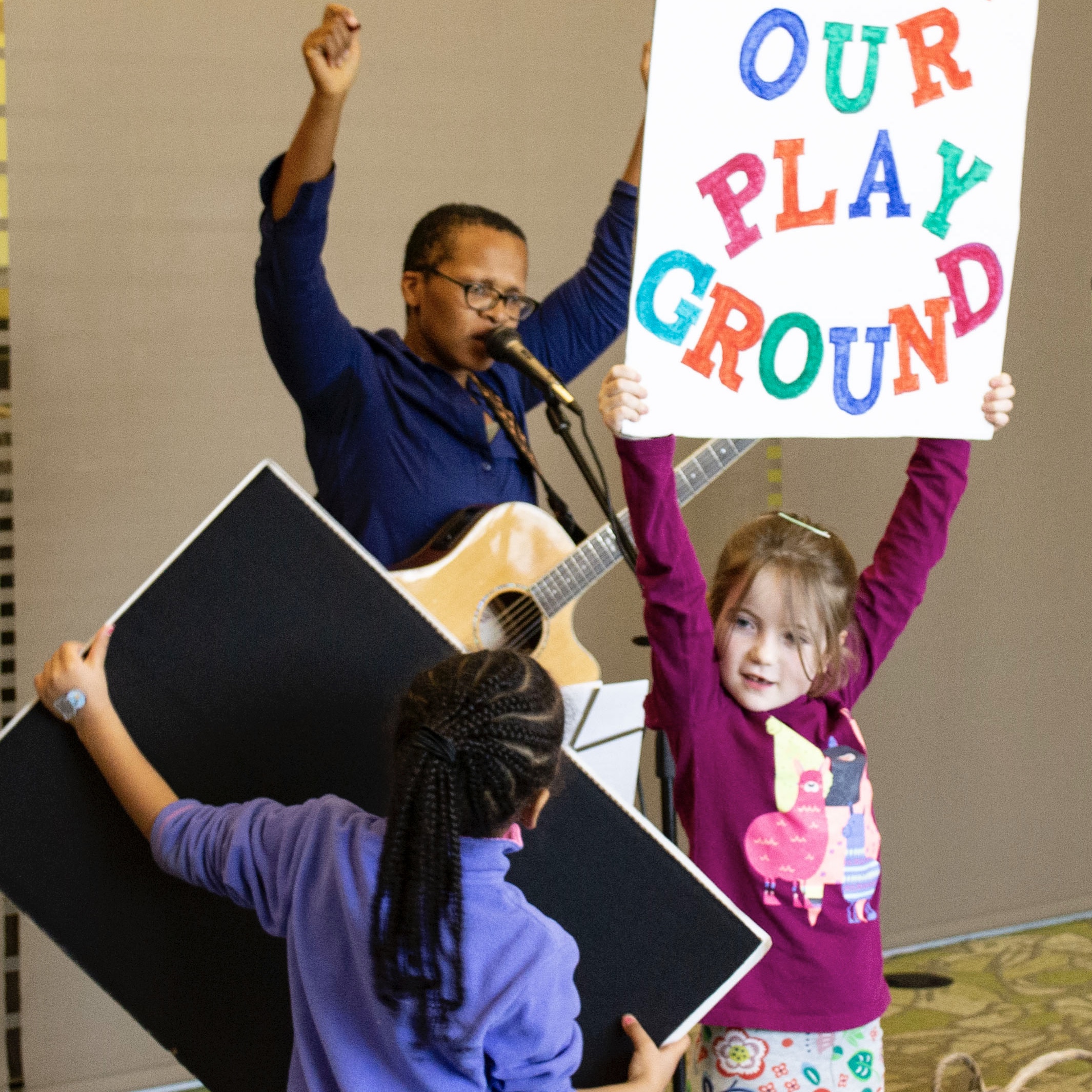 Lea Morris leads students in song. One student holds a sign that reads "Save Our Playground"