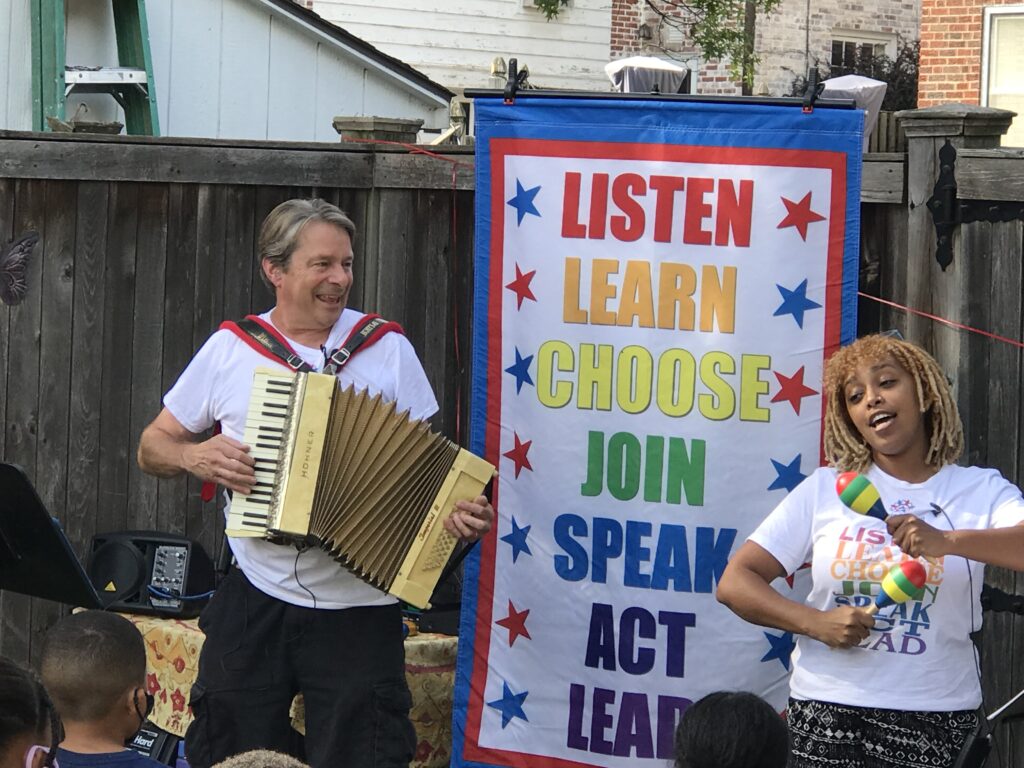 Teaching Artists leading students in song. A banner in the background reads: Listen, Learn, Choose, Join, Speak, Act, Lead.