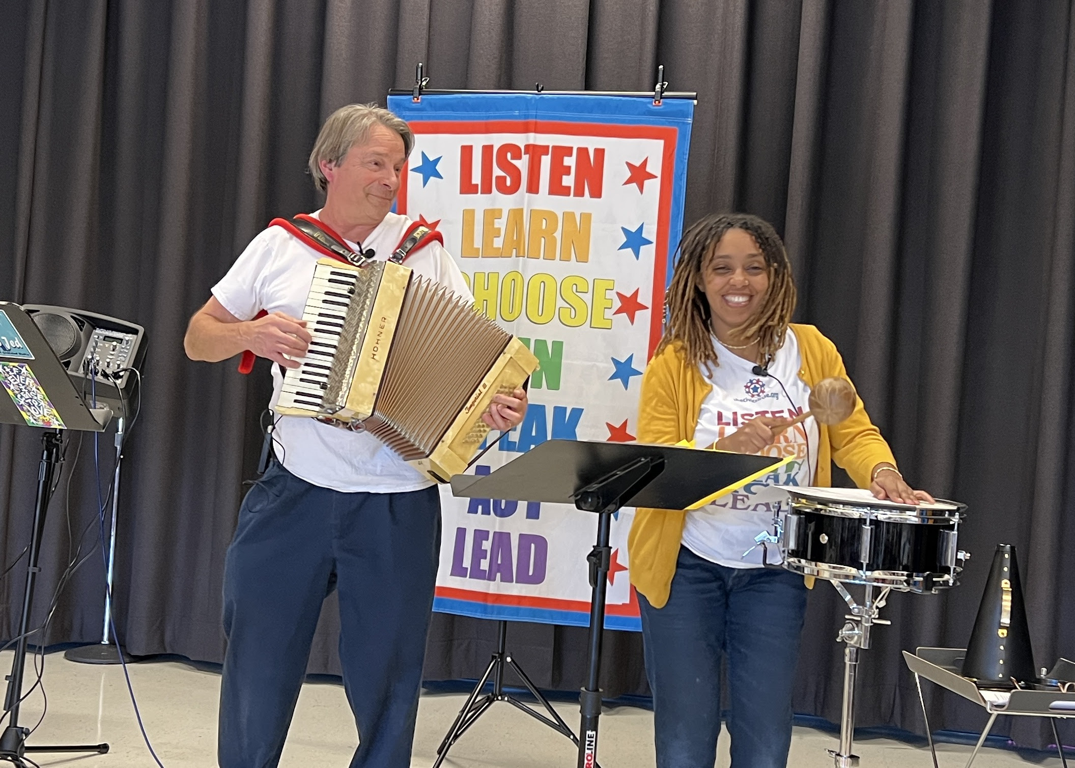 Nick Newlin and Munit Mesfin, smiling and playing musical instruments on stage. Behind them is a colorful poster that lists the seven steps to democracy (Listen, Learn, Choose, Join, Speak, Act, Lead).